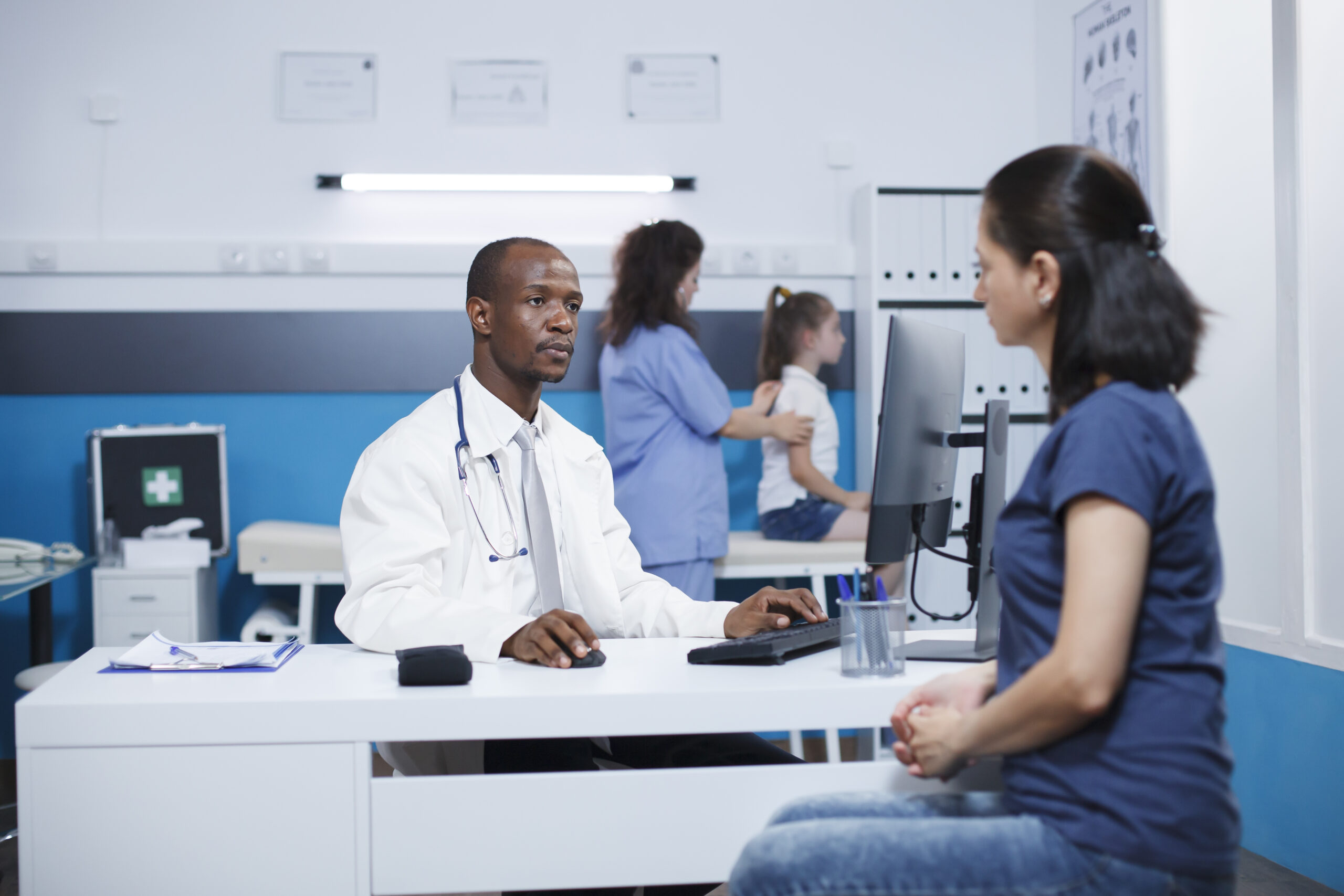 African American doctor in a lab coat explains treatment to a Caucasian woman in the hospital room. In the background, a female nurse does a medical examination on a young girl.
